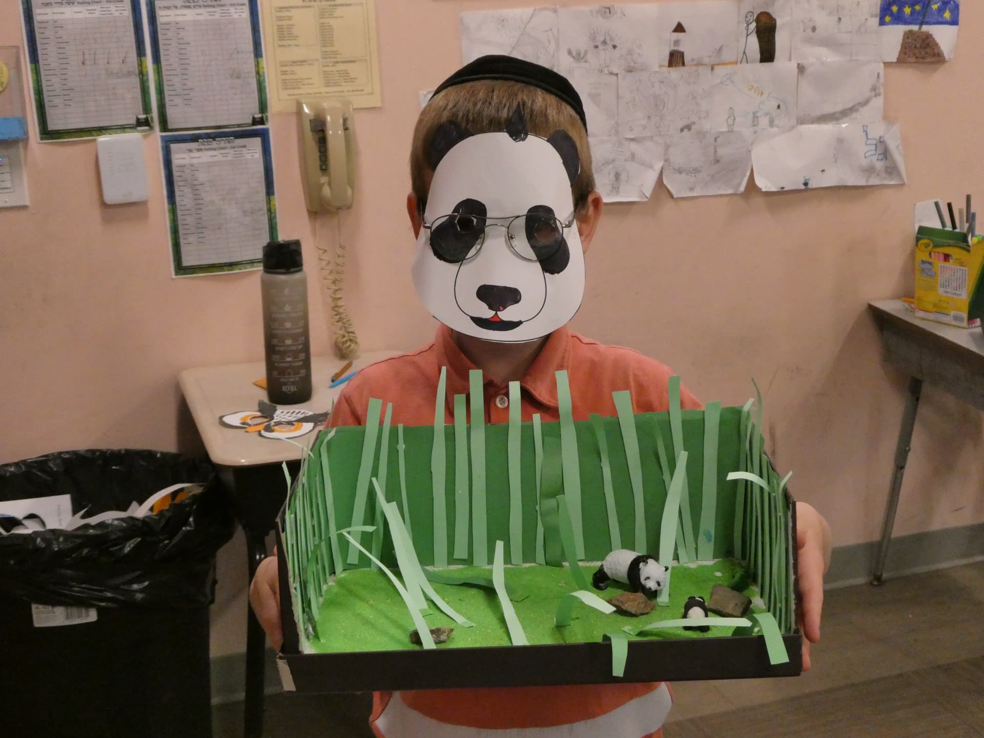 A student wearing a paper panda mask holding a handmade panda habitat diorama